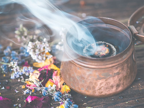 Ein kupfernes Gefäß verströmt Rauch, vermutlich von Räucherwerk oder Kräutern. Umgeben ist es von getrockneten Blüten und Blättern, darunter Lavendel. Die Szene wirkt meditativ, aromatisch und spirituell.